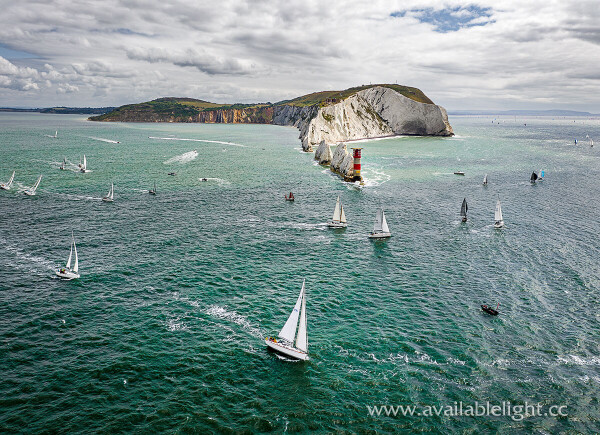 A drone shot of the coastline and a red and white striped lighthouse with yachts sailing around it. The sea is a mixture of blues, greys and turquoise. The boats mostly with white sails. An action shot, yet a restful image