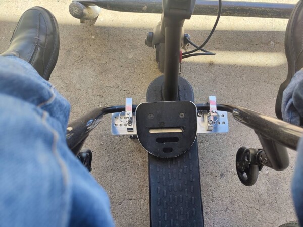 A view of how my electric scooter attaches to my wheelchair. The view is looking down, between my legs at the attachment. It has metal clamps my wheelchair's frame slides into with pins to hold it in place.