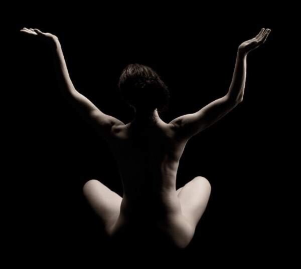 Rear view of me sitting cross legged and nude in chiaroscuro. Arms are upraised like a Minoan goddess. 