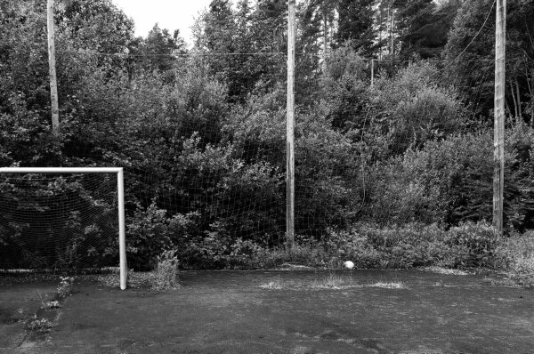 B&amp;W of half a soccer goal in front of a huge amount of bushes and trees. Also, the playing field has shrubs growing out of the cracks of the ground. White football is lying in the off-field beside the goal.