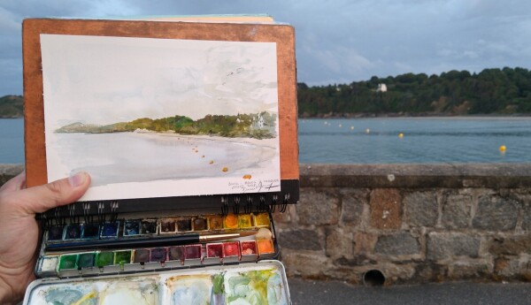 Photo of my hand holding a watercolor of a port , Binic in front of the landscape. The light changed while painting. I really liked the rythm of the little yellow floater on the sea.