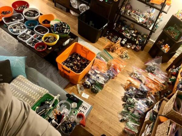 A living room with two large plastic bins that used to contains the LEGO collection of someone on its way to college. One bin is still half-full of loose parts, a few dozens of plastic bags labeled by LEGO set are lined up on the floor, while several plastic containers containing parts sorted by type are crammed on a coffee table.