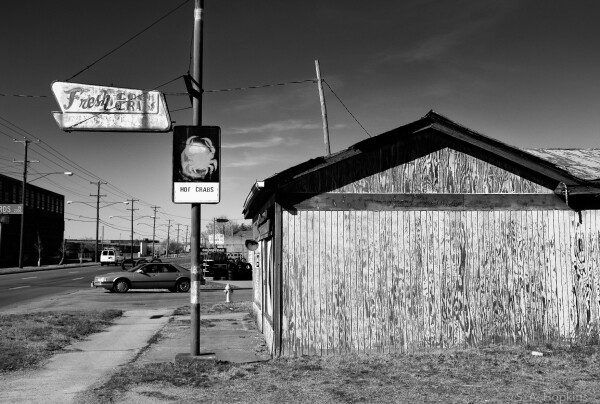 Black and white photo showing the side of a rustic old restaurant with one sign reading Hot Crabs and another, Fresh Crabs. The paint is peeling off of the side of wooden building. Judging by the state of decay, the business may have been closed for years. 