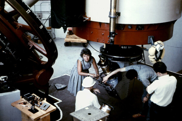 Color photo of Vera Rubin, perhaps in her 40s, working on a large apparatus with three men. She is looking at the camera, and wearing a gray-blue dress. 