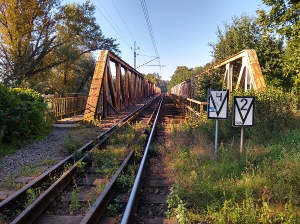 Zdjęcia z przodu mostu. Lewa strona jest zajęta torem kolejowym, z dodatkowymi szynami pośrodku i siecią trakcyjną u góry. Tor jest nieco zarośnięty roślinnością.

Po prawej stronie stoją dwa znaki, prostokątne, z malunkiem w kształcie litery V, i cyframi 4 i 2 odpowiednio w górnych częściach. Za nimi gęste krzewy odcinają dostęp do nieużywanej części mostu. Pośrodku widoczna jest barierka, a po bokach kratownice mostu.

Całkiem z lewej biegnie ścieżka do kładki dla pieszych. W pierwszej części ograniczona jest krzewami, a dalej barierką.