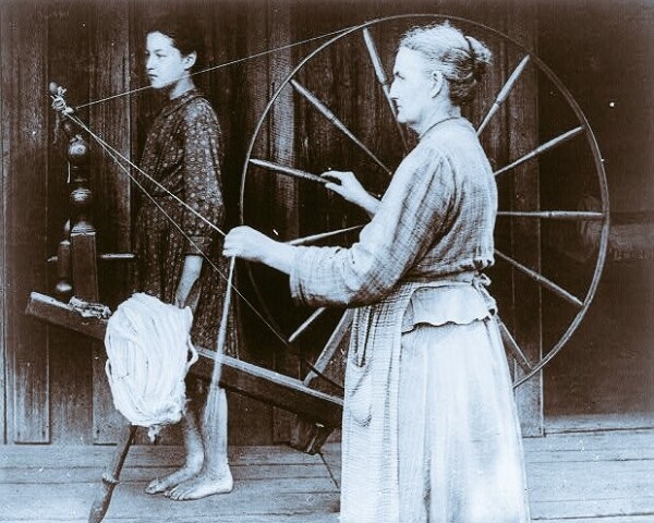 An unknown woman spinning with a wooden wheel, circa 1900.  Her hair is in a bun and she wears an apron over her long skirt.  A younger, barefoot woman, wearing a shorter dress, is standing in the background.