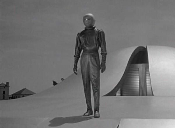 A scene from a black and white movie. It shows a huge, smooth UFO with a vertical opening in its top. A man dressed in a retro-future spacesuit and helmet has emerged. He's covered from head to toe, and his face isn't visible, so it's an eerie, though human-like, figure.