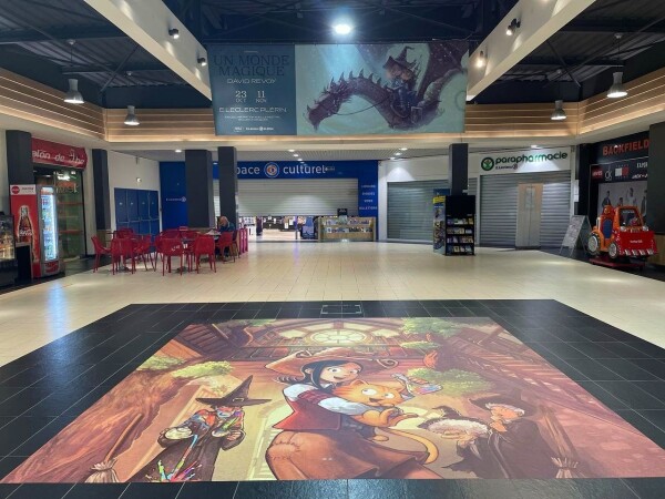 A photo of the hall of a supermarket with Pepper&Carrot art printed in big on the floor but also on a poster hanging from the ceilling. 