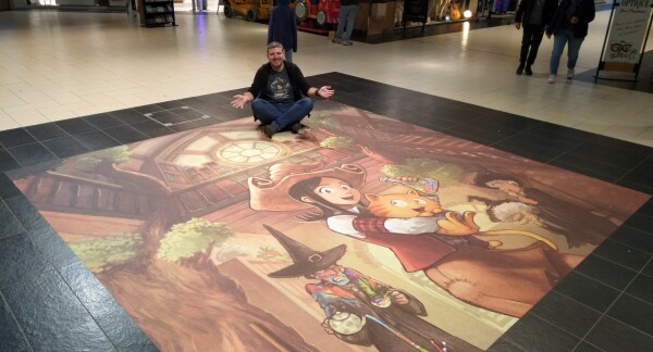 A photo at the mall in Leclerc Plérin France , I sit in a corner of my illustration printed on the floor. The cover book 2 of my webcomic Pepper and Carrot.