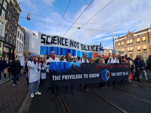 Scientists in white lab coats walk through the streets of Amsterdam carrying banners with the texts "Science not silence" and "Scientist Rebellion. The privilege to know, the duty to act."