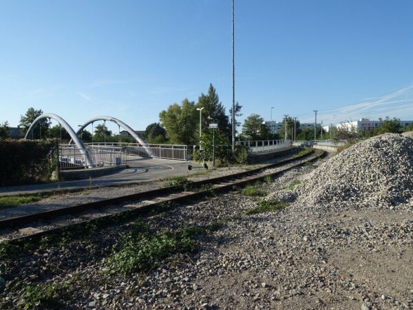 Bogenförmiges Industriebahngleis durch Schotterfläche bzw. dahinter auf eine Brücke übergehend, rechts davon ein Schotterhaufen, links davon ein Rad- und Fußweg, der um eine Kurve auf eine weitere Brücke mit bogenförmiger Architektur Richtung linken Bildrand abbiegt