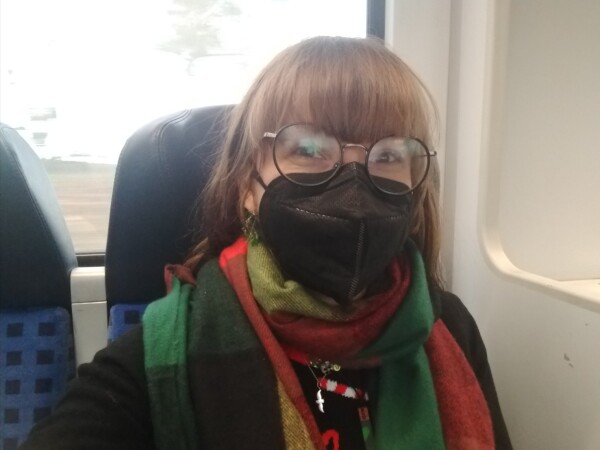 Ich mit schwarzer Maske, rotbrauner Ponyfrisur und großer runder Brille und bunten Schal auf einem blauen Sitz am Fenster im Regionalzug.