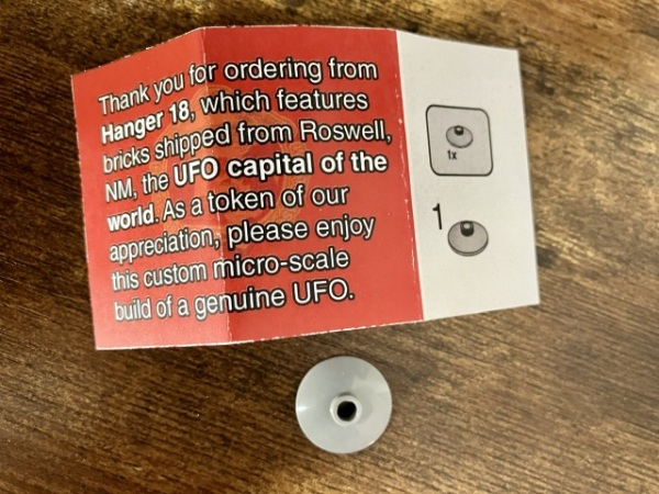 A small piece of paper on a desk reads “Thank you for ordering from Hanger (sic) 18, which features bricks shipped from Roswell, NM, the UFO capital of the world. As a token of our appreciation, please enjoy this custom micro-scale build of a genuine UFO.” Next to the text, a one-step one-part LEGO instruction shows a small gray dish. Next to the paper on the desk, here is the expected small gray dish part. 