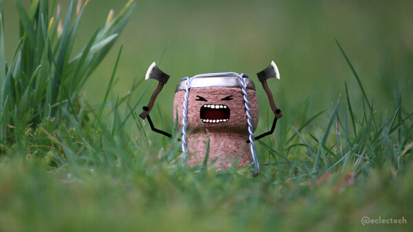 Close up photo of a cork from a bottle of prosecco, with it's metal cap and wire cage in place, sitting in a grassy lawn. I have drawn on a shouty face, and arms wielding axes, so it looks like a warrior going into battle.