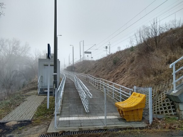 Rampa z płyt chodnikowych, idąca pod górę. Z przodu krótki płot, po bokach barierki. Po lewej stronie tabliczka, za nią budynek techniczny. Wzdłuż chodnika lampy uliczne. Na nasypie widać znak z nazwą przystanku.

Nie ma już drzew. Podpory nasypu zastąpiono betonem z barierką.