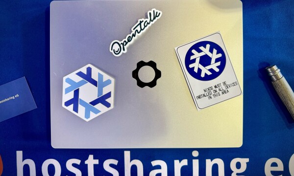 silver framework laptop on blue ground, white big letters that read „hostsharing eg“ - one unattached sticker on the laptop with the nixos logo, one with a blue variant and text that reads upon quote. one opentalk logo sticker also unattached, a gray wooden folded opinel knife on the right