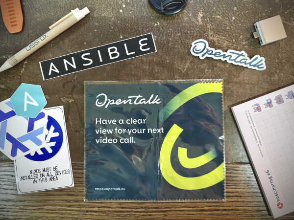 background is an old wooden, leddery desk, a big square opentalk themed microfiber cloth is in the bottom center of the image. it reads below the OpenTalk lettering "Have a clear view for your next video call." - on the left there are two NixOS stickers, one reads "NixOS must be installed on all devices in this area". On the two stickers is also one octagon shaped Ansible logo sticker. Next to them is an Ansible signature sticker where the A is the Ansible logo. Further above is a paperly looking pen, branded "Osssrox". On the right is an OpenTalk lettering sticker, a Framework Laptop USB module laying around and a Hostsharing eG checkered notepad with variants of their mascot decorating its right side. There is also a Fuju lenshood laying around for fun and a piece of Garmin watch bracelet