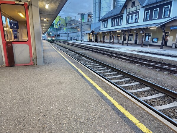 Bahnsteig in Kirchdorf/Krems. Ein Passagierzug fährt in den Bahnhof ein, gezogen von einer ÖBB 1044 Lok. Im Hintergrund das Kirchdorfer Zementwerk.