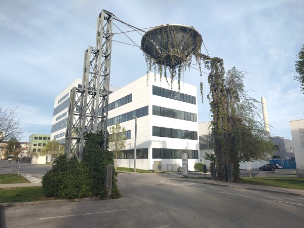 Eine Zufehrtsstraße mit mehstöckigem weißem Bürogebäde dahinter. Über der Straße hängt ein großes Trichterförmiges Gebilde an einem Gestänge, das auf Stehern an beiden Straßenseiten befestigt ist, die gesamte Konstruktion ist aus Metall. Goße Teile davon sind mit grünen Schlingpflanzen bewachsen.