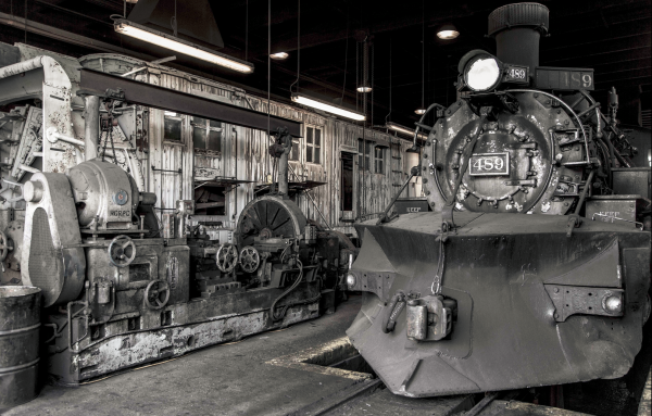 B&W 3/4 view of a narrow-gauge steam locomotive with a snowplow on its pilot beam, behind which is an enormous lathe.