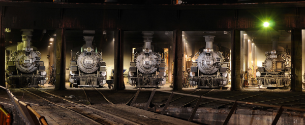 Night view of the fronts of various narrow gauge steam locomotives in the Durango roundhouse, with the turntable visible in front of them