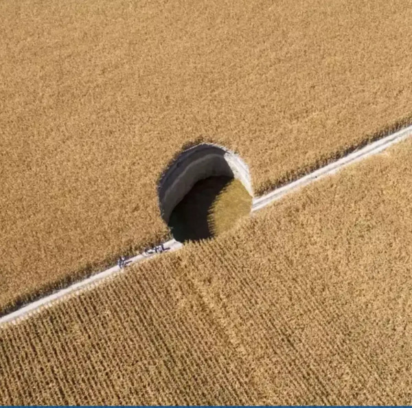 A wheat field and a road with a 25 meter almost perfect circle diameter and 8 meters deep hole in the middle, people like small dots looking from a distance.