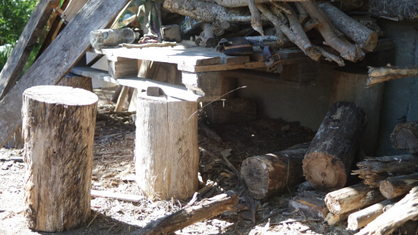 Eine Holzpalette auf ein paar Holzstrünken unter dem Vordach auf der Terrasse. Auf der Palette ist Holz, unter ihr Reste von Holz, Sand und Schotter.