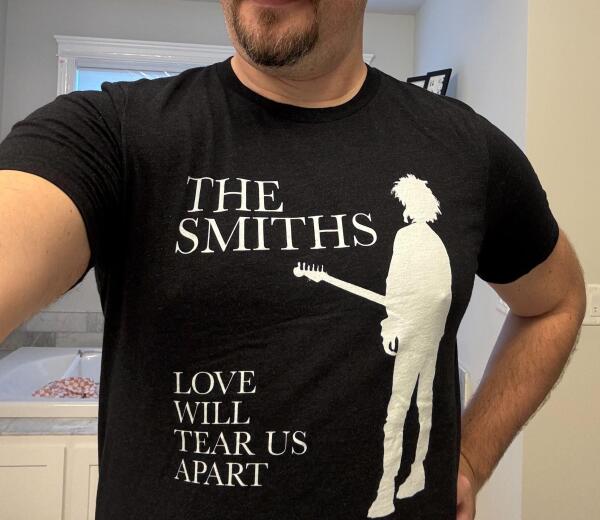 a black shirt featuring a white silhouette of Robert Smith holding a guitar. Large text reads “The Smiths” and “Love Will Tear Us Apart”