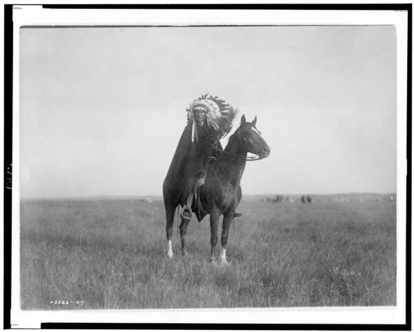 The image depicts a figure of an individual who appears to be wearing Native American attire, specifically what is known as a headdress or traditional headwear. The person is mounted on a horse and seems poised in the midst of movement, possibly galloping through an open field characterized by sparse vegetation under a cloudy sky.

In terms of historical context, this photo was taken during Edward S. Curtis's time documenting Native American life, which is suggested by his name tag as indicated below it. This photograph appears to be from "The Prairie Chief," published in 1907 according to the caption found at its bottom right corner.