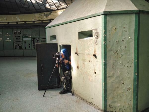 A photographer with blue hair and camouflage pants is taking a photo with a tripod from inside an old bunker in a green control room