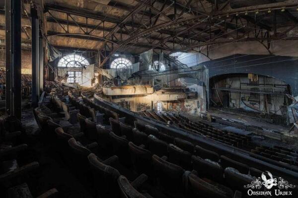 A wide view of a decaying, abandoned theater with rows of seats and a stage. The theater has large arched windows