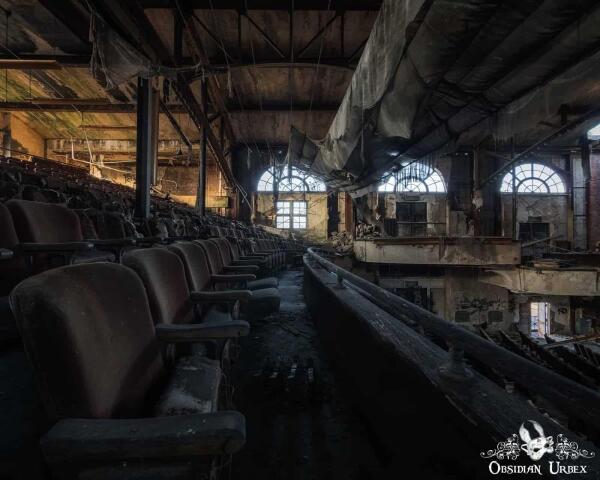 An abandoned theatre shows rows of empty seats, dilapidated curtains, and broken windows. 