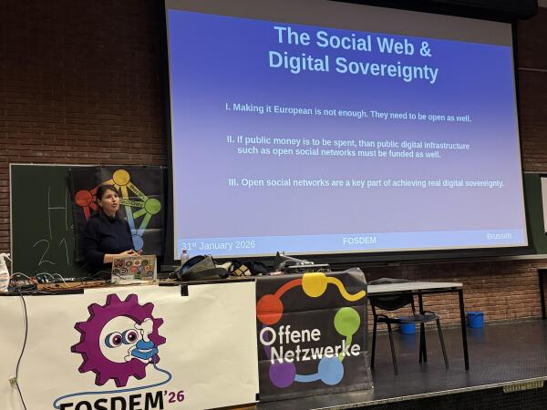 A woman is presenting at a conference called FOSDEM, standing in front of a screen displaying a presentation titled "The Social Web & Digital Sovereignty." Key points outline the importance of open digital infrastructure and funding open social networks.
