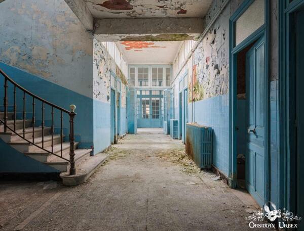 An abandoned French thermal spa corridor with peeling blue paint, a staircase, and natural light from windows ahead.