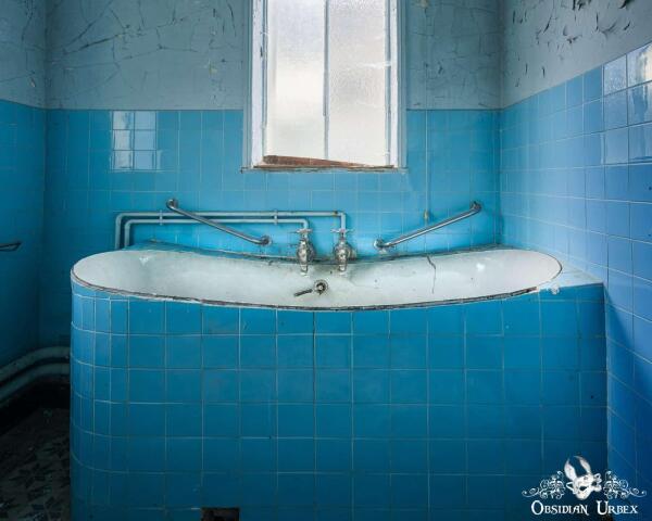 An abandoned, blue-tiled treatment room features a curved white bathtub and window. The room shows signs of decay.