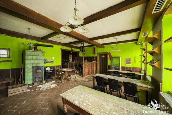 An abandoned inn's dining area features vibrant green walls, exposed wooden beams, and dirty tables.