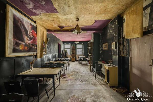 A long, abandoned restaurant interior shows a decaying room with dark walls, peeling purple and yellow ceiling, and scattered tables and chairs.