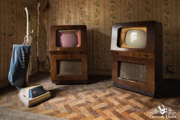 An abandoned room features a vintage blue Hoover vacuum and two old wooden televisions on a patterned floor with floral wallpaper.