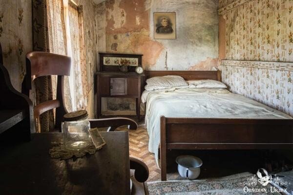 A decaying, abandoned British bedroom featuring peeling wallpaper, a simple bed with white sheets, and a framed portrait.