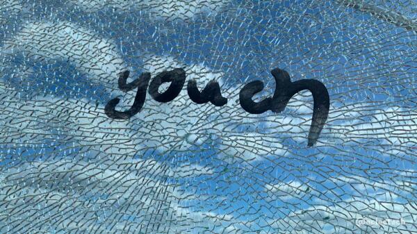 A photo taken through a smashed panel of glass. The glass must be reinforced in some was as it has stayed together very well despite shattering. Behind the pattern of broken glass you can see a blue sky with a lot of fluffy white clouds. On top of the glass someone has painted "youch" in black paint just above the central hole of the shatter pattern.