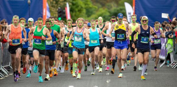 Runners at the start of a mass participation road race, with green facial recognition detection boxes overlaid on their faces and race bib numbers, illustrating automated biometric identification in a public sporting event.