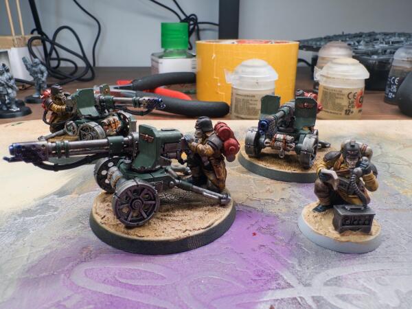 The image features several detailed miniature models of soldiers with flamethrower cannons and soldiers engaged in various roles. The setting includes painting supplies and tools, with a focus on the intricately designed figures positioned on a textured surface.