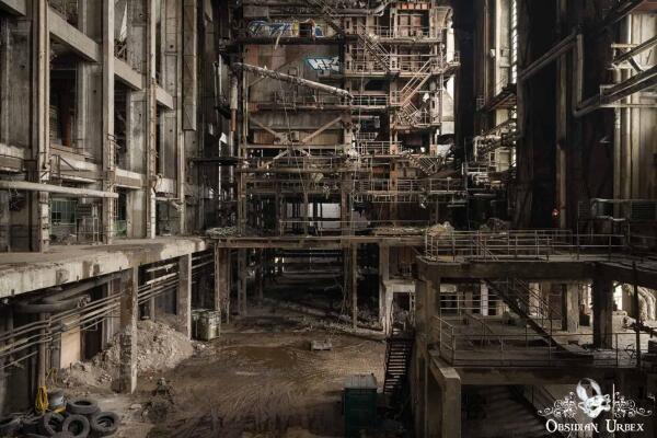 An expansive, desolate view inside an abandoned industrial building, with multi-level structures, pipes, and debris