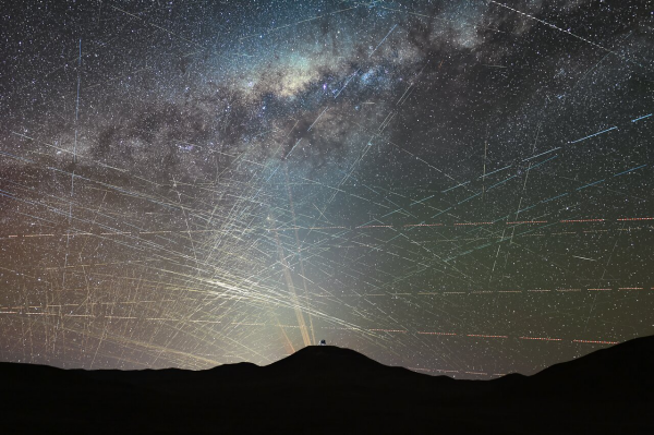 The background of this image shows a starry sky, including the bright lane of the Milky Way. Hundreds of bright streaks cross the sky in different directions, as if scratching the background natural sky behind it. The bottom of the image is occupied by the dark silhouette of a mountainous desert landscape. Atop the mountain at the centre, is a small, distant metallic dome and yellow-orange lasers shine behind it.