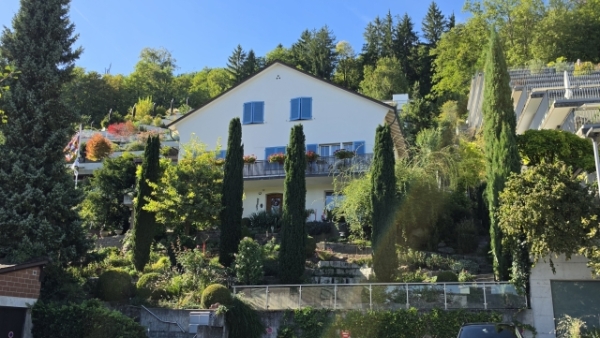 Ein helles Wohnhaus mit blauen Fensterläden und Balkon steht an einem dicht bepflanzten Hang mit Bäumen und Sträuchern.