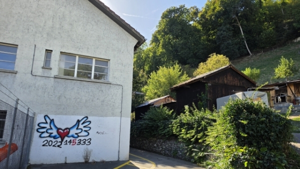 An einer hellen Gebäudewand ist ein Graffito mit einem geflügelten roten Herz und der Aufschrift „2022 145333“ zu sehen; daneben stehen dunkle Holzbauten und dichter Bewuchs.