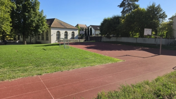 Eine rote Laufbahn umgibt eine Rasenfläche; daneben befindet sich ein rotes Spielfeld mit Basketballkörben und einem niedrigen Gebäude.