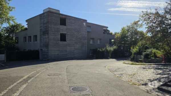 Ein modernes graues Gebäude mit einem holzverkleideten Gebäudeteil steht an einer Straßenkreuzung zwischen Bäumen.
