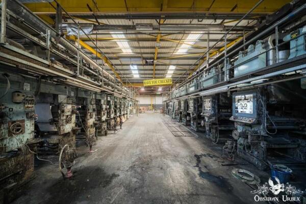 An abandoned wallpaper factory hall features rows of large, rusty printing machines