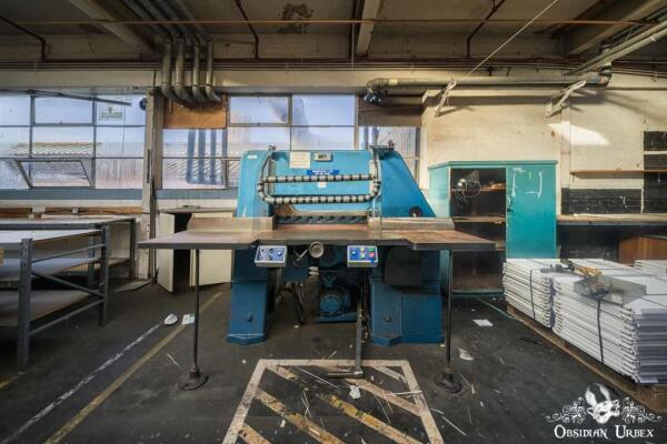 A large blue industrial cutting machine stands central in an abandoned factory, with stacks of material nearby